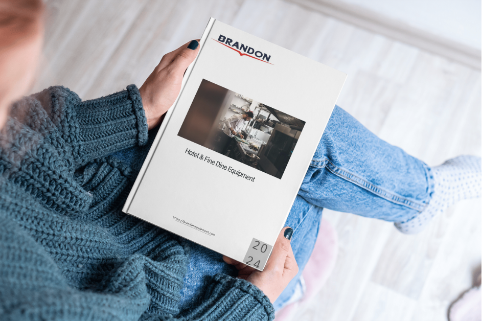 The image shows a person sitting comfortably in a relaxed environment, holding a catalog. The cover of the catalog prominently features the "BRANDON" logo at the top, with the title "Hotel & Fine Dine Equipment" below a blurred image of a kitchen scene. The year "2024" is displayed in the bottom right corner. The person is wearing a cozy blue sweater, and the overall scene suggests a calm and comfortable moment of browsing through the catalog. The catalog cover design is clean and professional, suggesting high-quality content inside.